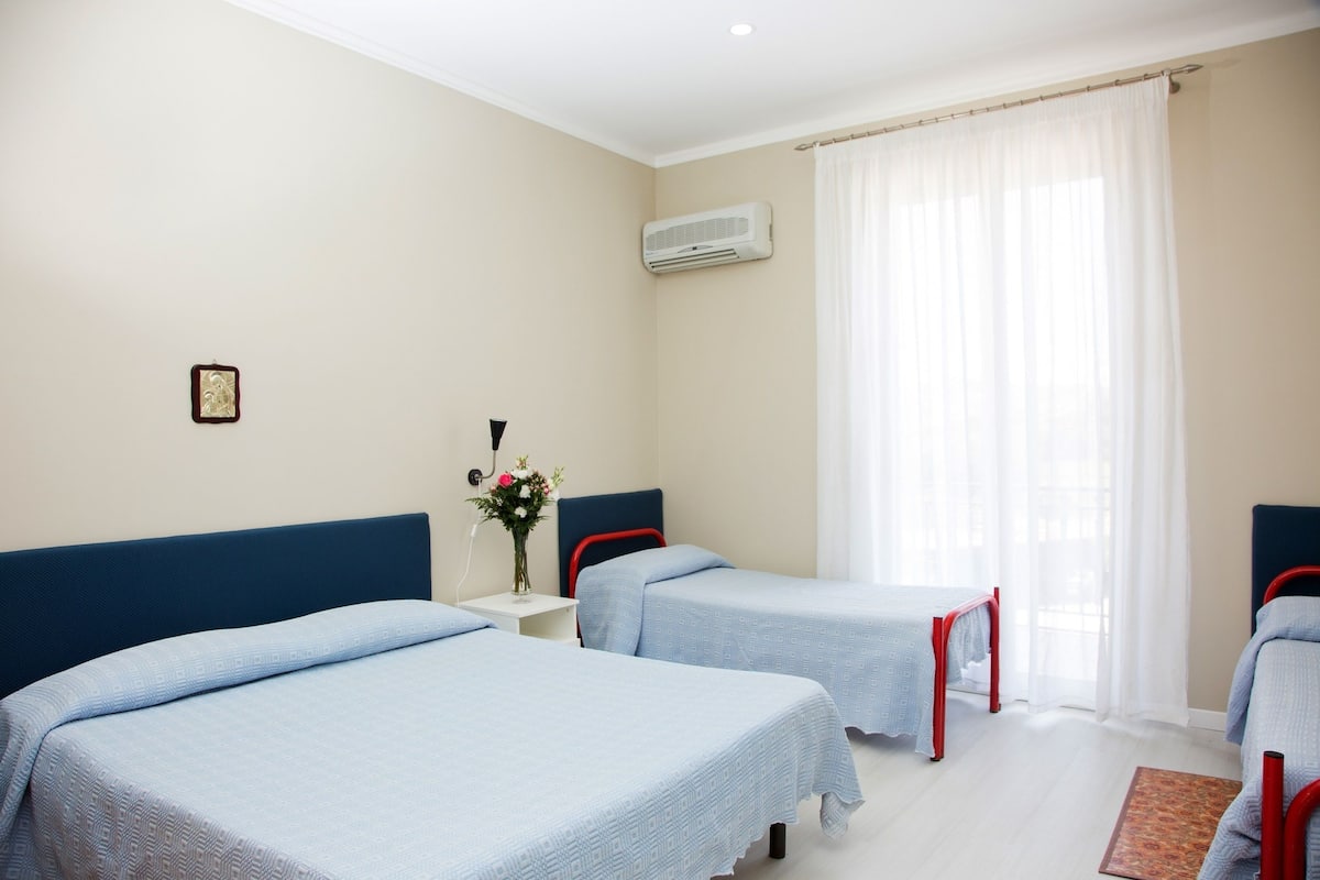 A hotel room with two beds, a small bedside table with a vase of flowers, an air conditioning unit, and a window with sheer curtains.