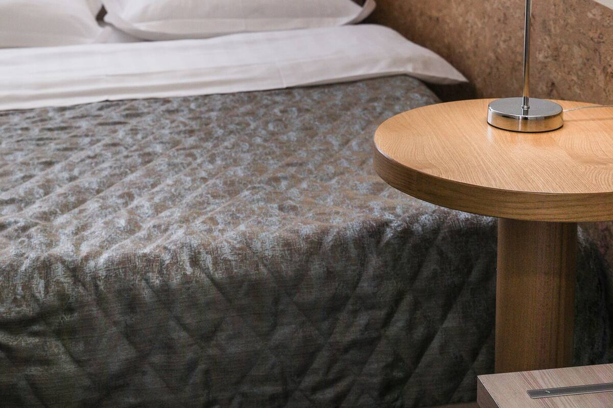 Hotellihuone, jossa on sänky, pyöreä puinen pöytä, jossa on lamppu, ja seinälle asennettu valaisin.