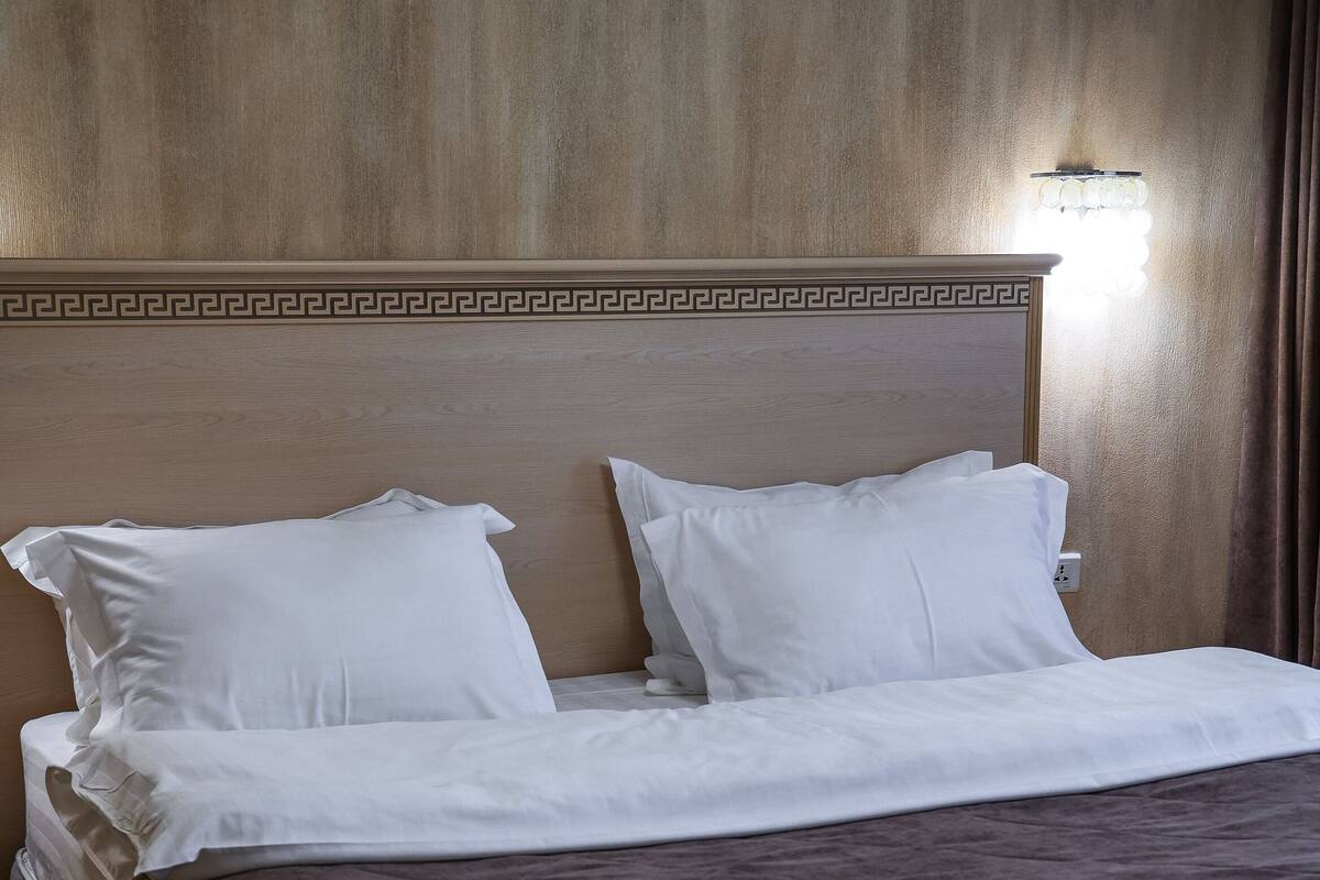 Siististi pedattu sänky valkoisilla lakanoiden ja ruskealla peitteellä varustetussa hotellihuoneessa, jossa on puinen pääty ja seinään kiinnitetty valaisin.