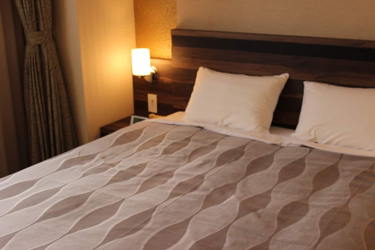 Una habitación de hotel con cama, mesita de noche, lámpara y cortina.