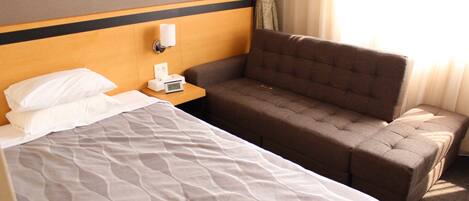 A hotel room with a bed, a sofa, a nightstand with a lamp, and a window with curtains.
