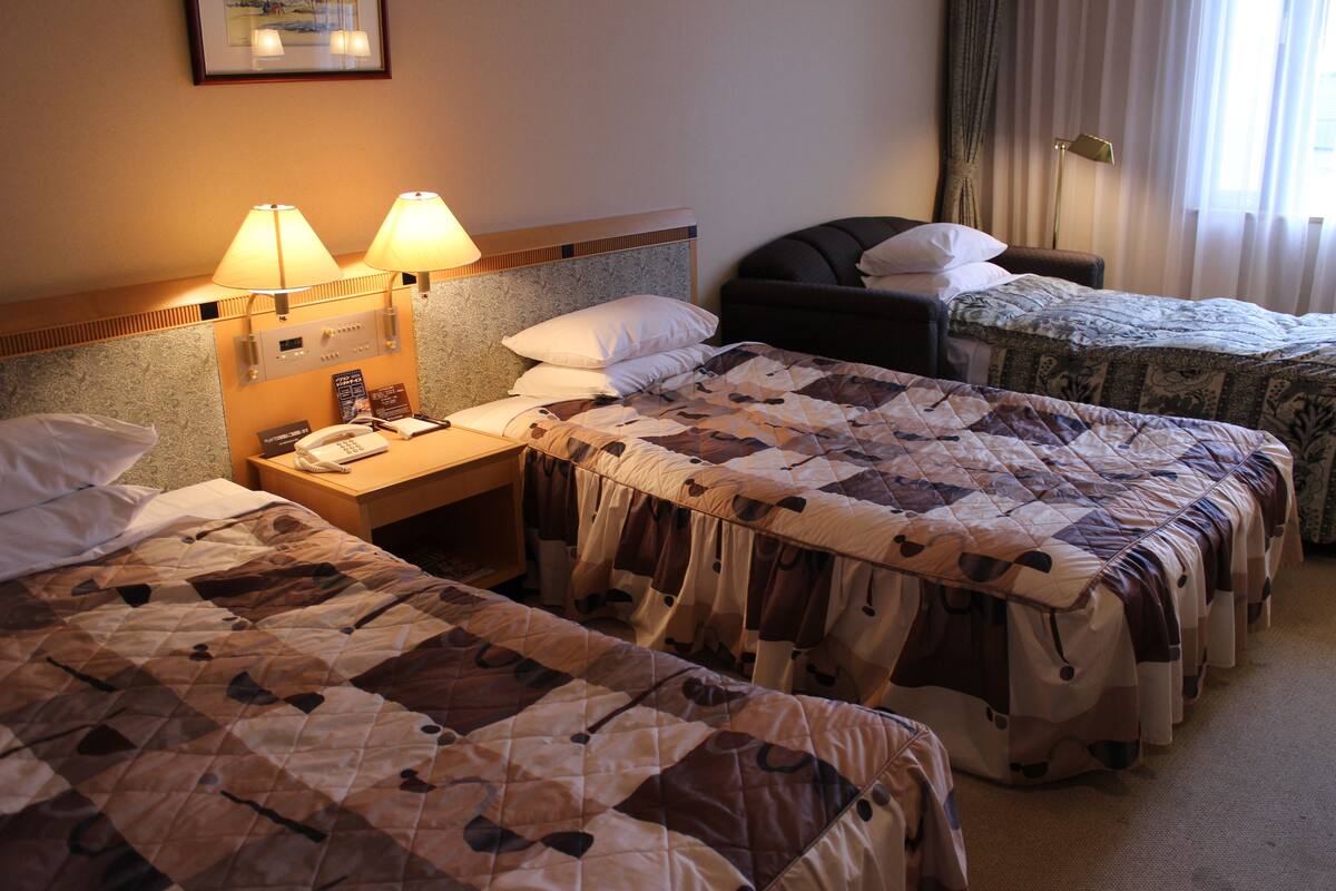 Habitación de hotel con dos camas individuales, una mesita de noche con lámpara y teléfono, una silla y una ventana con cortinas.