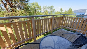 Une terrasse en bois avec une table et des chaises en verre, donnant sur une étendue d’eau et des arbres.