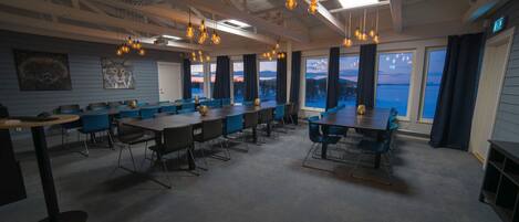 Un salón de conferencias con varias mesas y sillas, amplias ventanas con vista a un lago y lámparas colgantes.