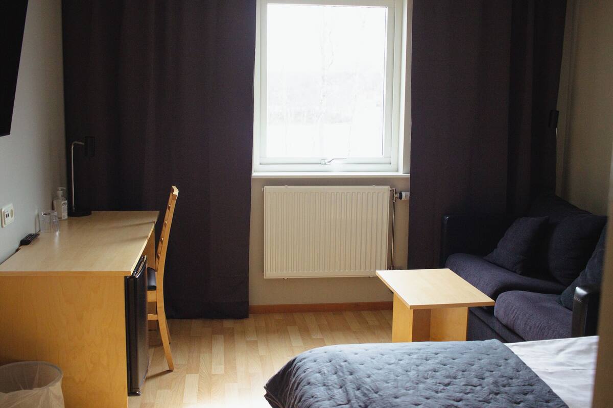 Pieni huone, jossa on sänky, pieni työpöytä, tuoli, pöytä ja ikkuna, jossa on verhot.