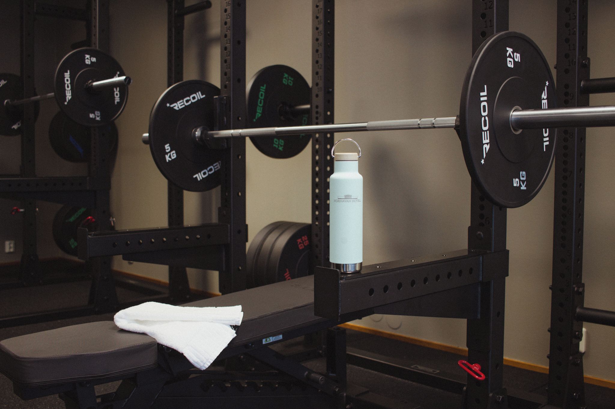 A weightlifting bench with a barbell loaded with weight plates, a water bottle, and a towel.