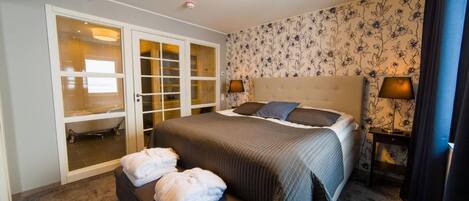 Une chambre à coucher avec un lit, deux lampes de chevet, un banc avec des serviettes, une salle de bain fermée par des parois de verre et un papier peint à motifs floraux.