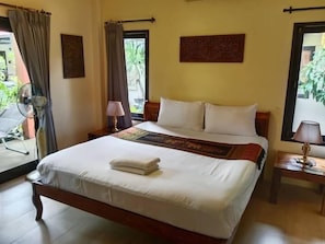 A neatly made bed with a white comforter and a folded towel, a bedside lamp, a small table with a lamp, and a window with curtains. - Baan Tropicana (Koh Samui)