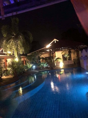 A nighttime swimming pool area with a curved design, surrounded by well-lit plants and a building with a visible entrance. - Baan Tropicana (Koh Samui)
