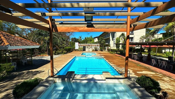 Una piscina con una pérgola encima, rodeada de mobiliario exterior y vegetación.