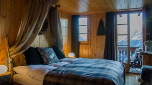 A wooden cabin bedroom with a bed, curtains, a nightstand, and a view of a snowy mountain. - Hotel Le Grand Chalet Favre (Anniviers)