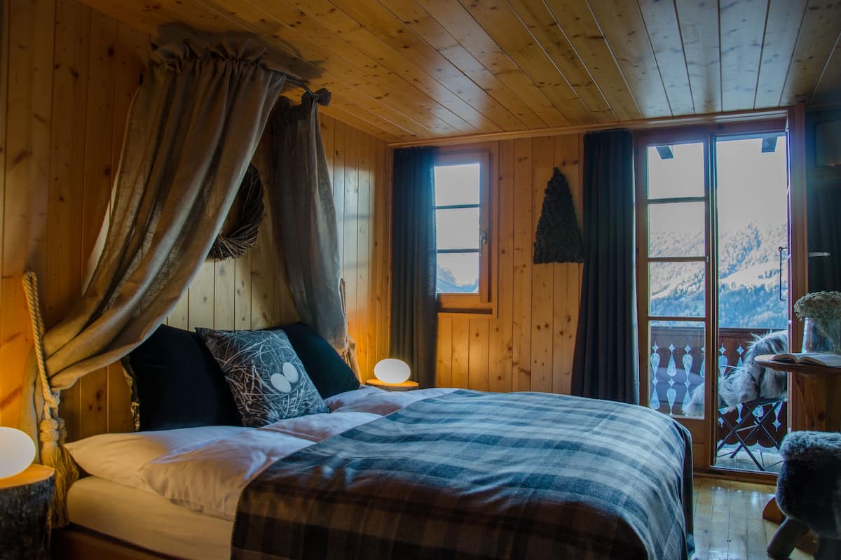 A wooden cabin bedroom with a bed, curtains, a nightstand, and a view of a snowy mountain.