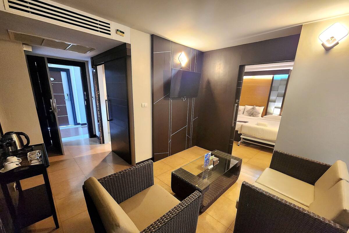 Una habitación de hotel moderna con mobiliario de mimbre, una mesa de centro de vidrio y una cama con edredón blanco.