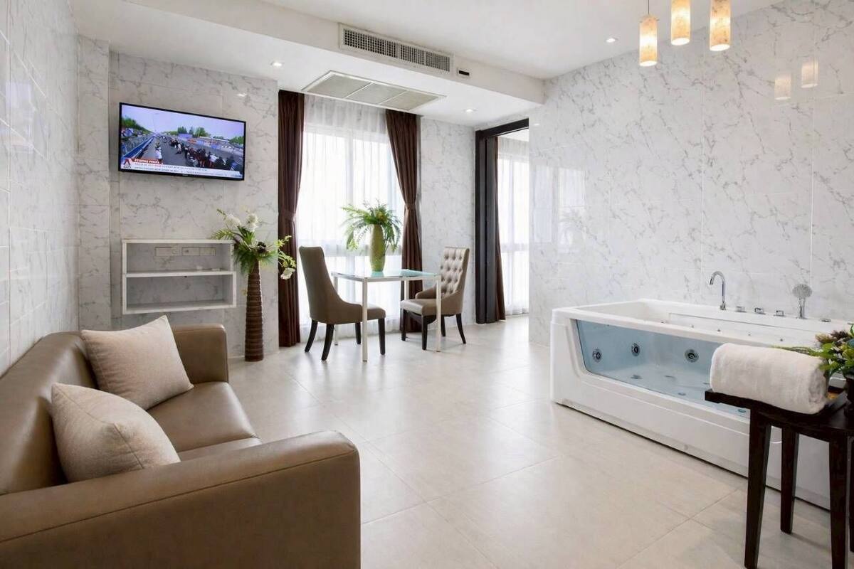 Una habitación de hotel moderna con sofá, una mesa de comedor con sillas, televisor, una bañera grande y una planta en maceta.