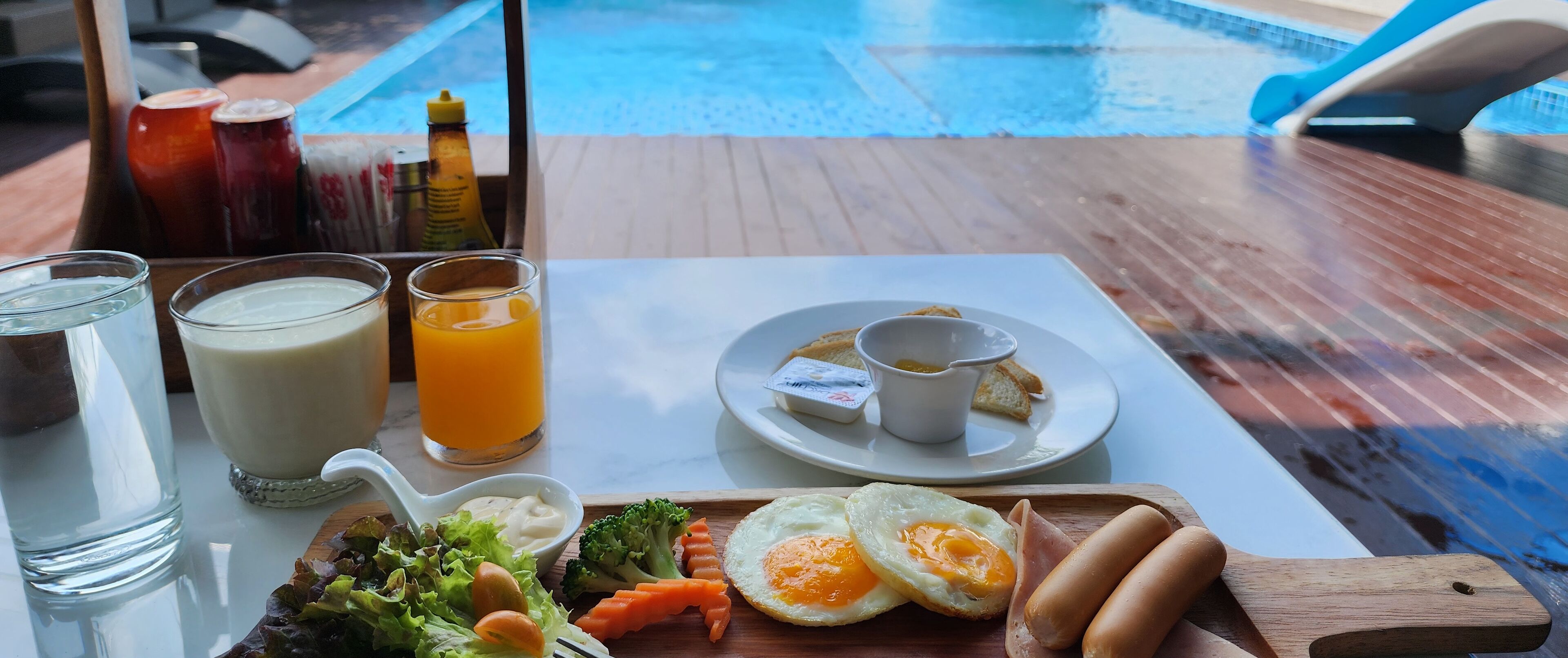 A breakfast tray with eggs, sausages, toast, and a side salad, along with two glasses of milk and orange juice, and condiments on a table by a pool.