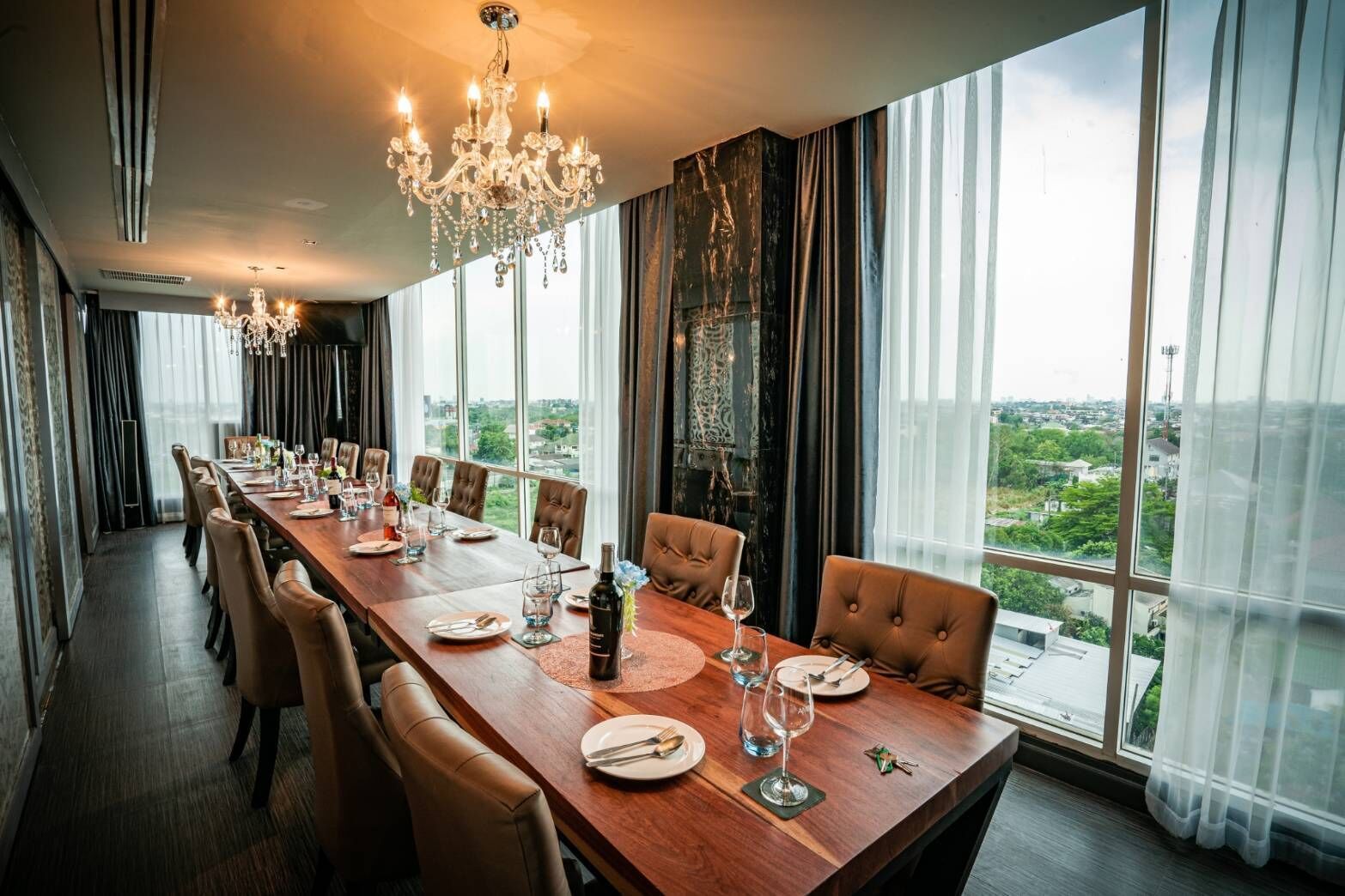 A dining room with a long wooden table set for a meal, large windows offering a city view, and a chandelier above.