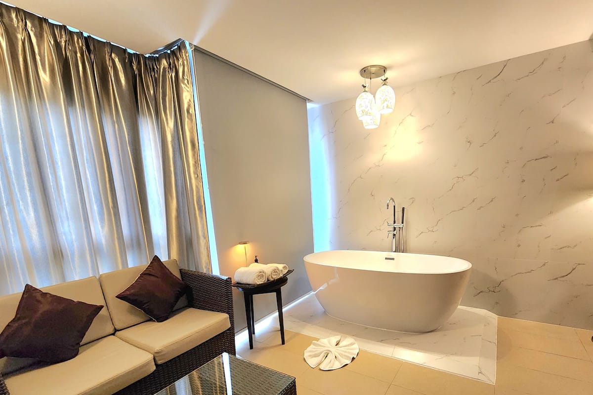 Un baño moderno con una bañera independiente, un sofá con cojines, una mesita de noche con una lámpara y un gran ventanal con cortinas.