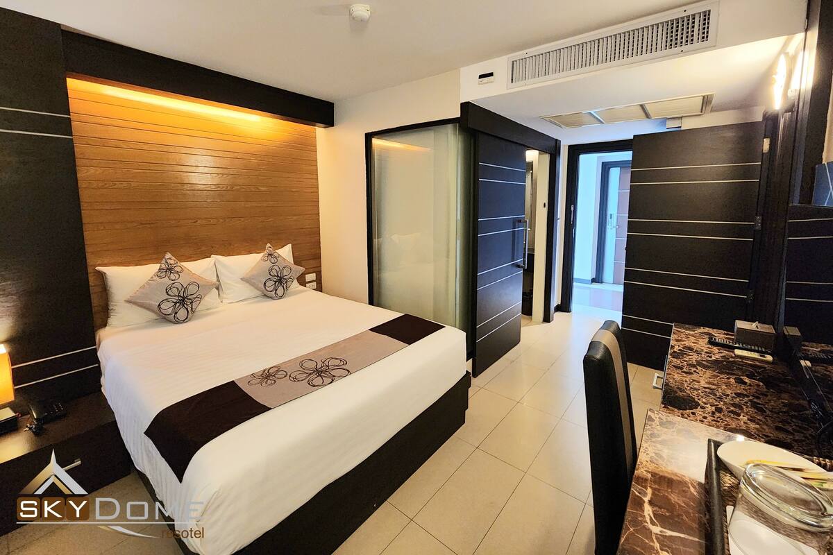 Una habitación de hotel moderna con una cama grande, un cabecero de madera, un separador de vidrio y una encimera de mármol con un lavabo.
