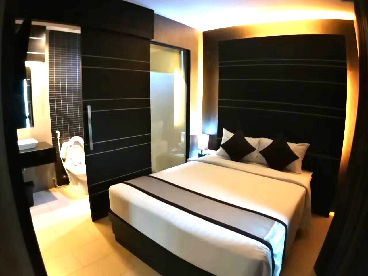 Una habitación de hotel moderna con una cama grande, una mesita de noche, un espejo y un baño con inodoro y lavamanos.