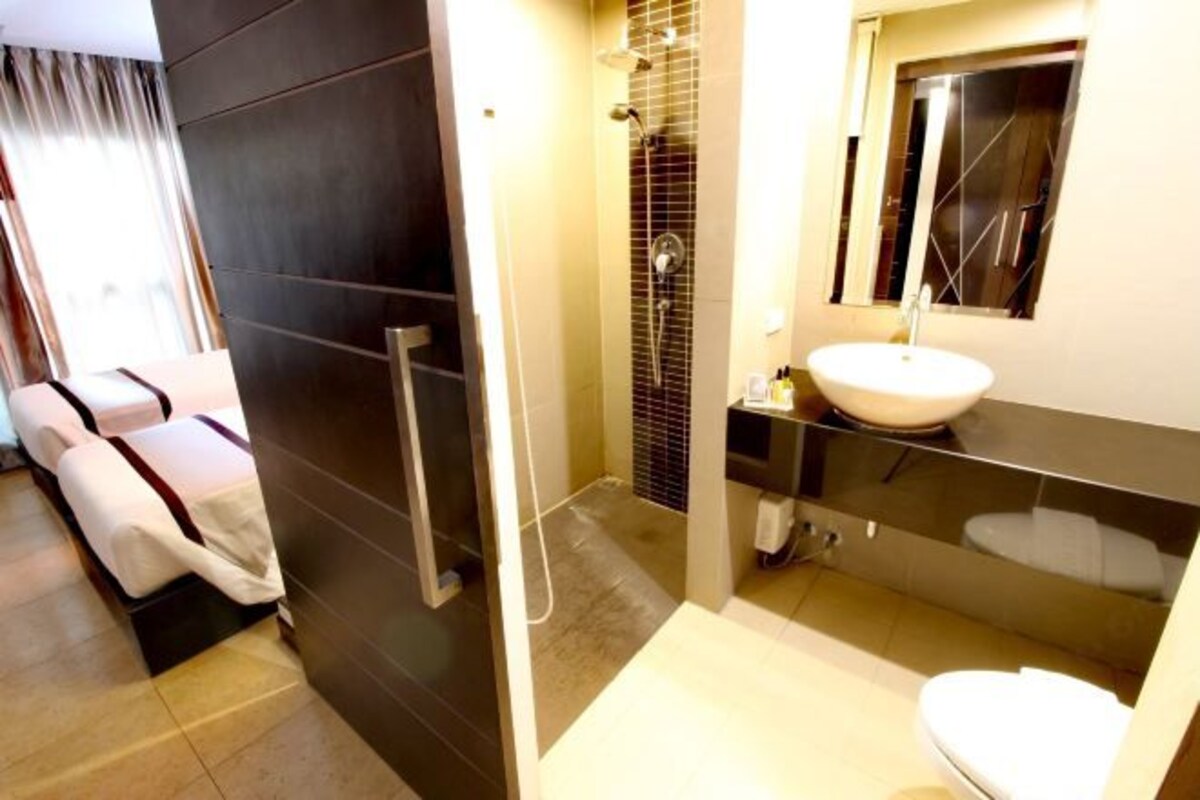 A modern bathroom with a shower, a sink, and a mirror. There is a bed with white linens in the adjacent room.