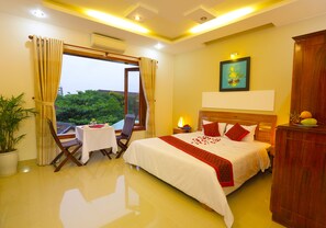 A hotel room with a bed, a table, a chair, a potted plant, and a view of greenery outside.