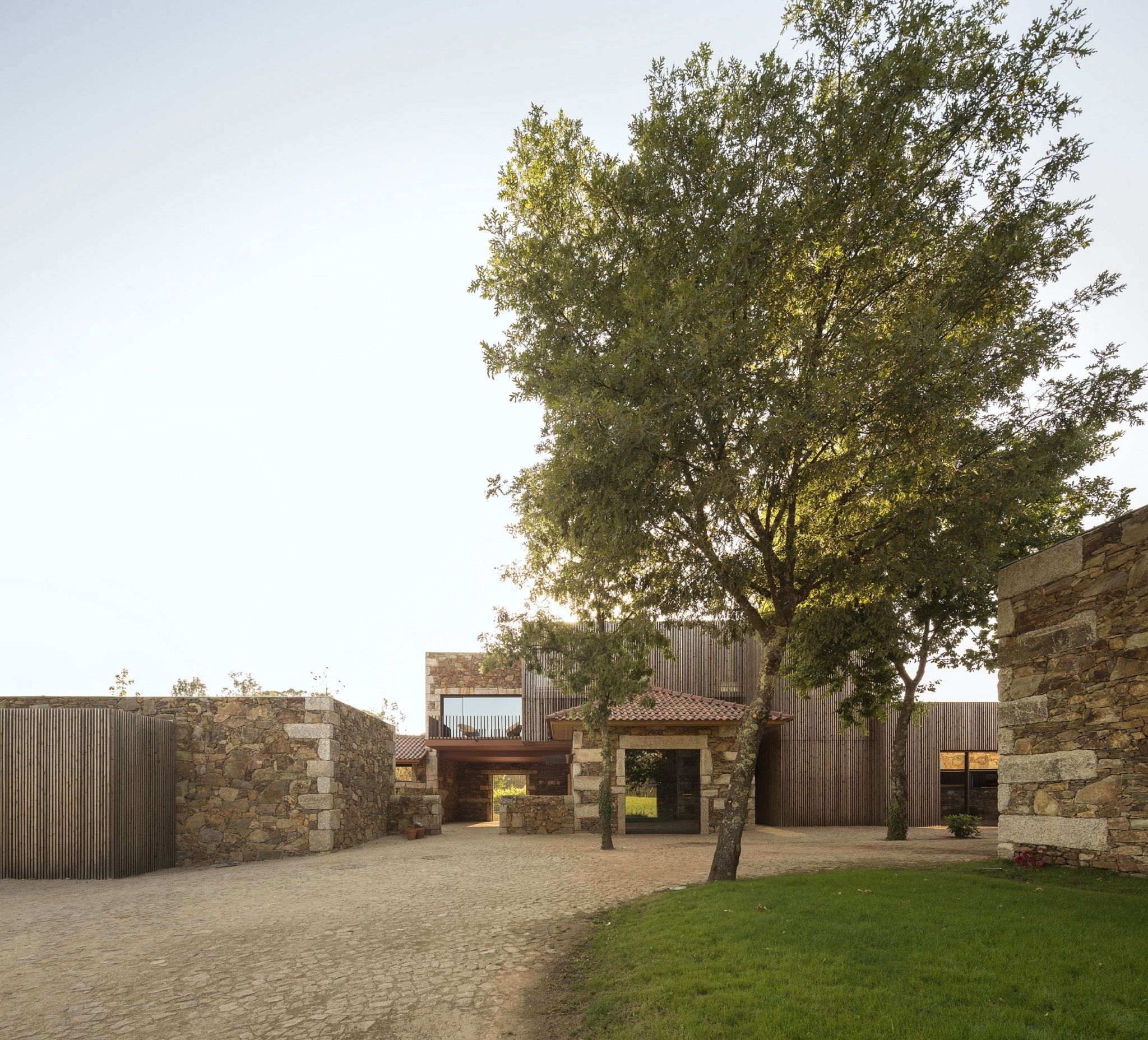 A courtyard with a stone wall, a tree, and a modern building with a tiled roof.
