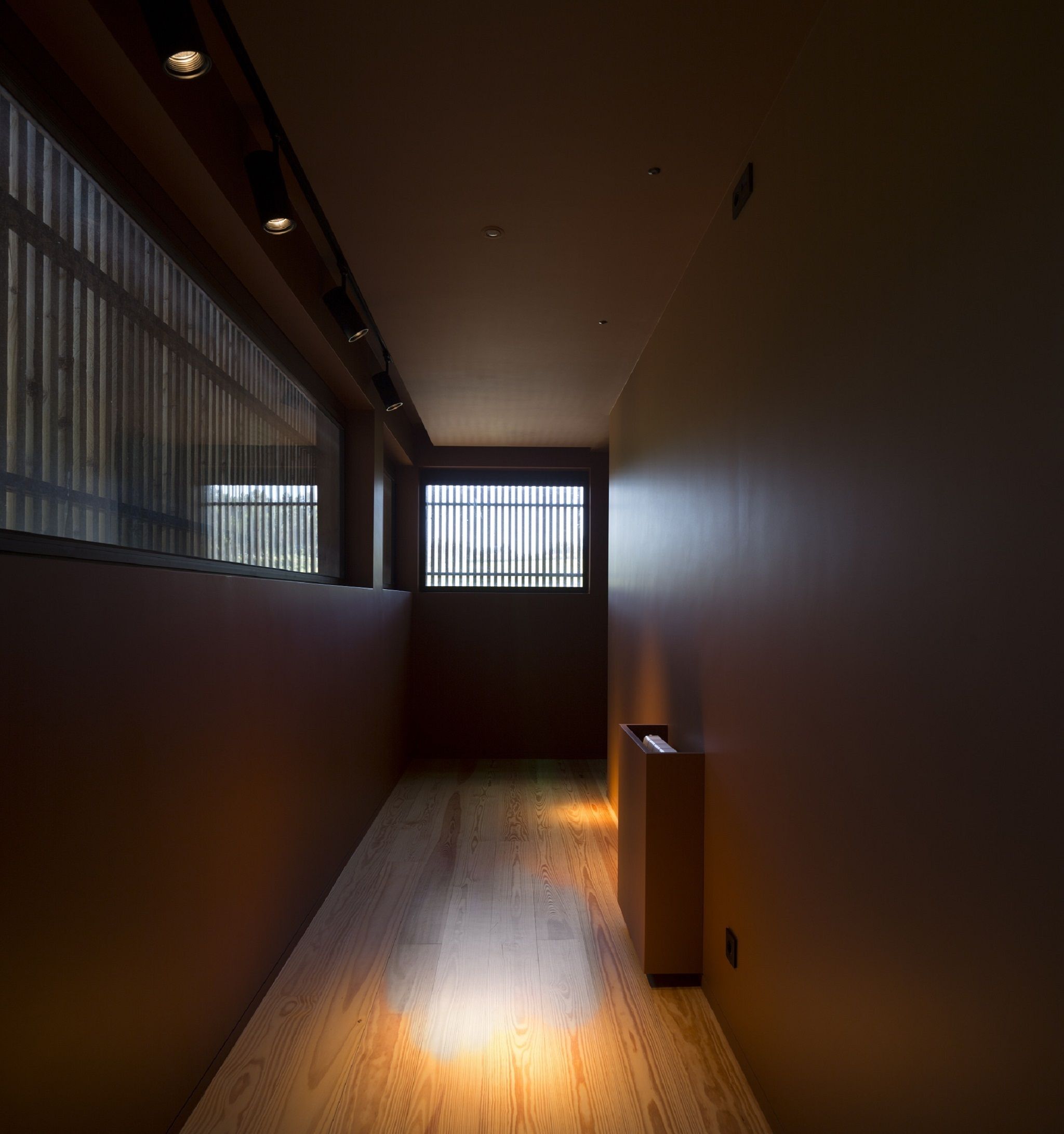 A narrow hallway with wooden flooring and a window with vertical blinds.