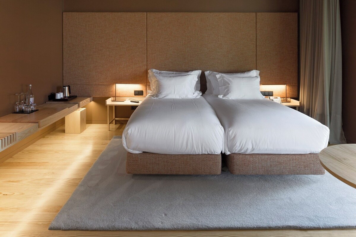 A modern hotel room with a large bed, bedside tables, a desk, and a large window with curtains.