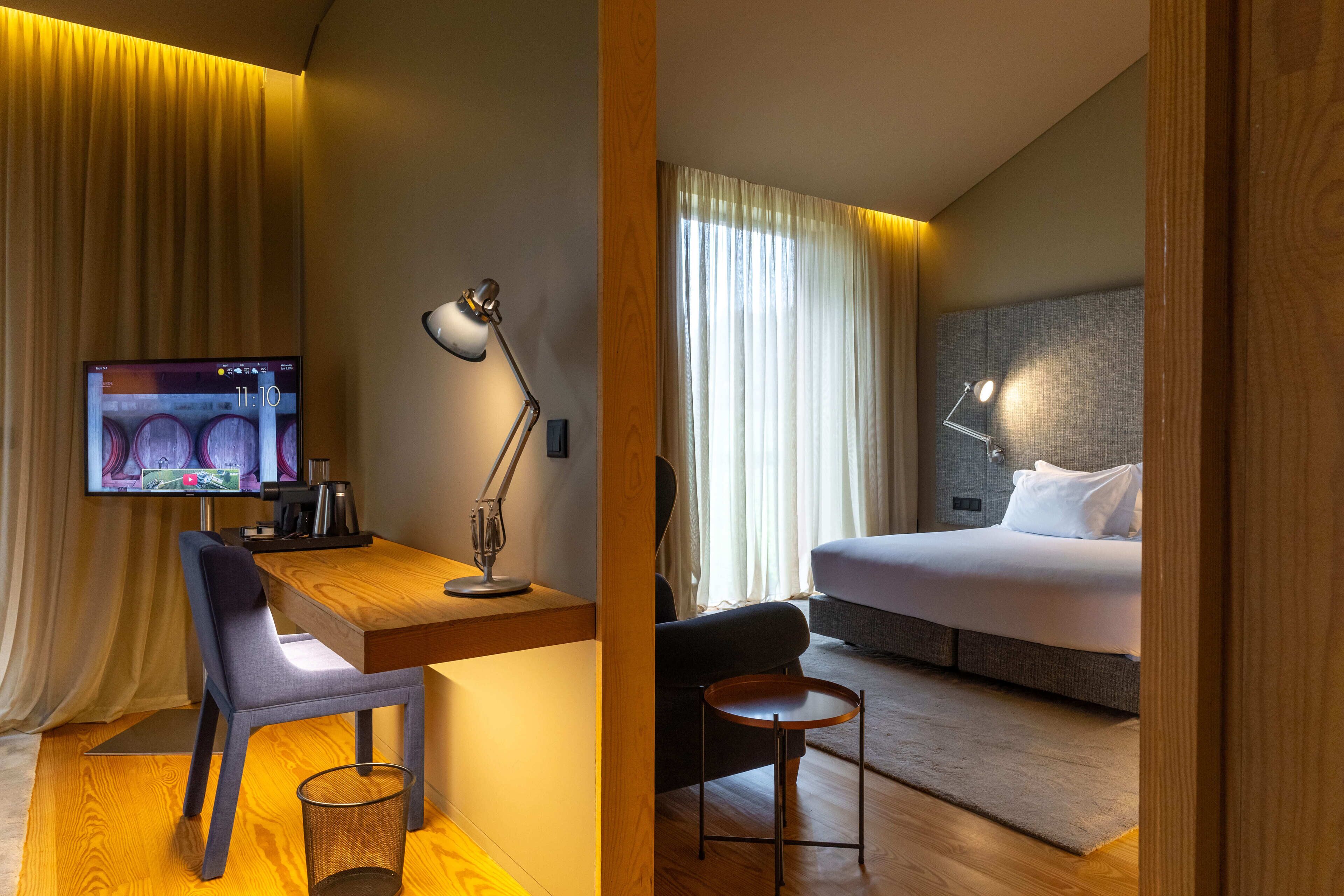 A hotel room with a wooden desk, a chair, a television, a bed, and a nightstand.