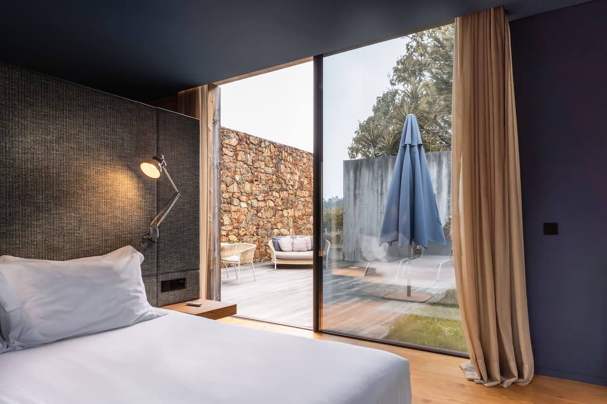 A modern bedroom with a large bed, a desk, a lamp, and a view of an outdoor patio with a stone wall and an umbrella.