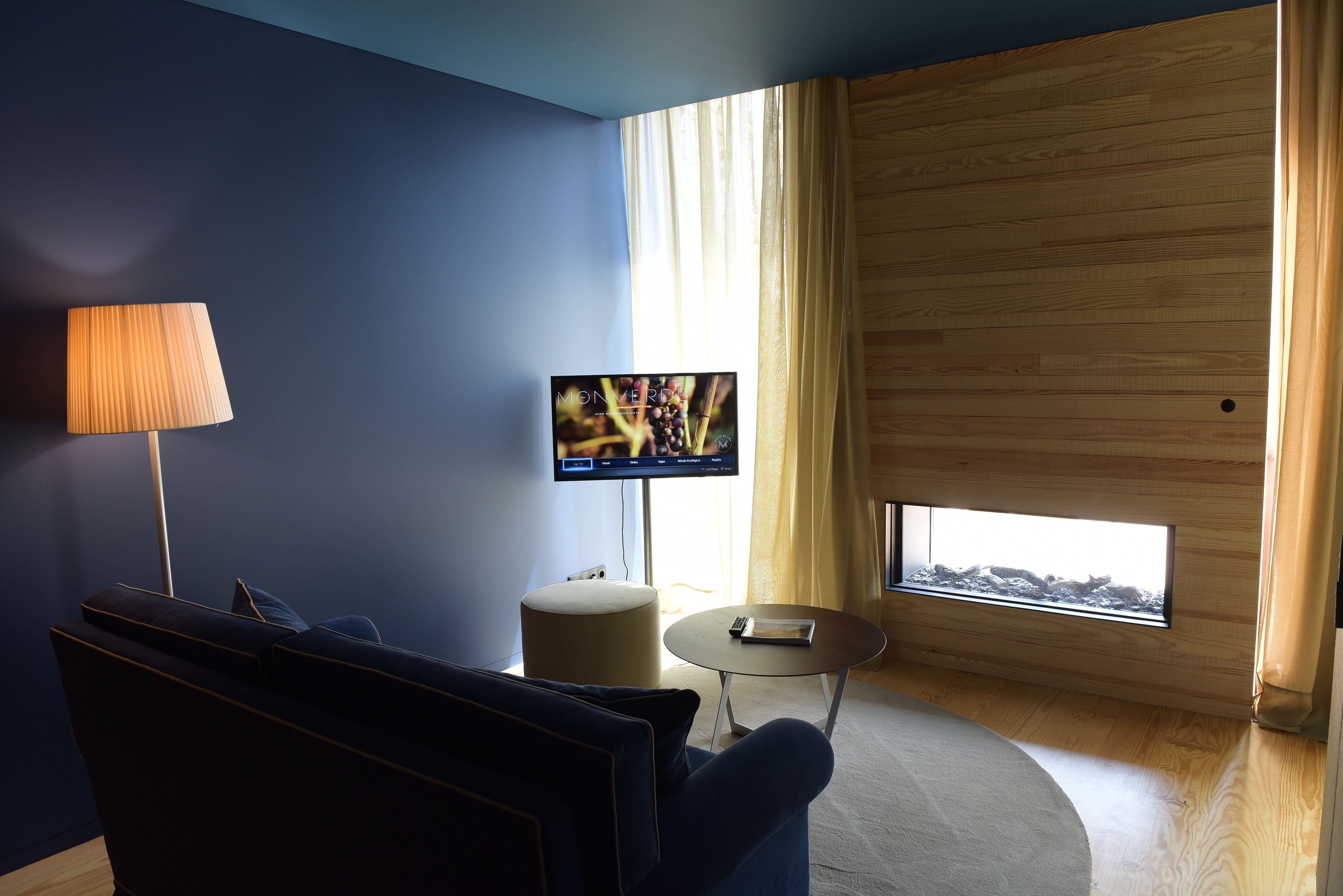 Una sala de estar moderna con una pared azul oscuro, piso de madera, un sofá, una mesa de centro redonda, una lámpara y un televisor que muestra una escena de la naturaleza.