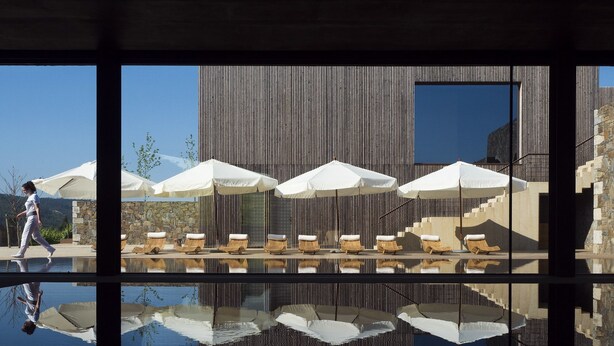 Un edificio moderno con una piscina que refleja su estructura. Hay sillones de descanso y sombrillas cerca de la piscina.