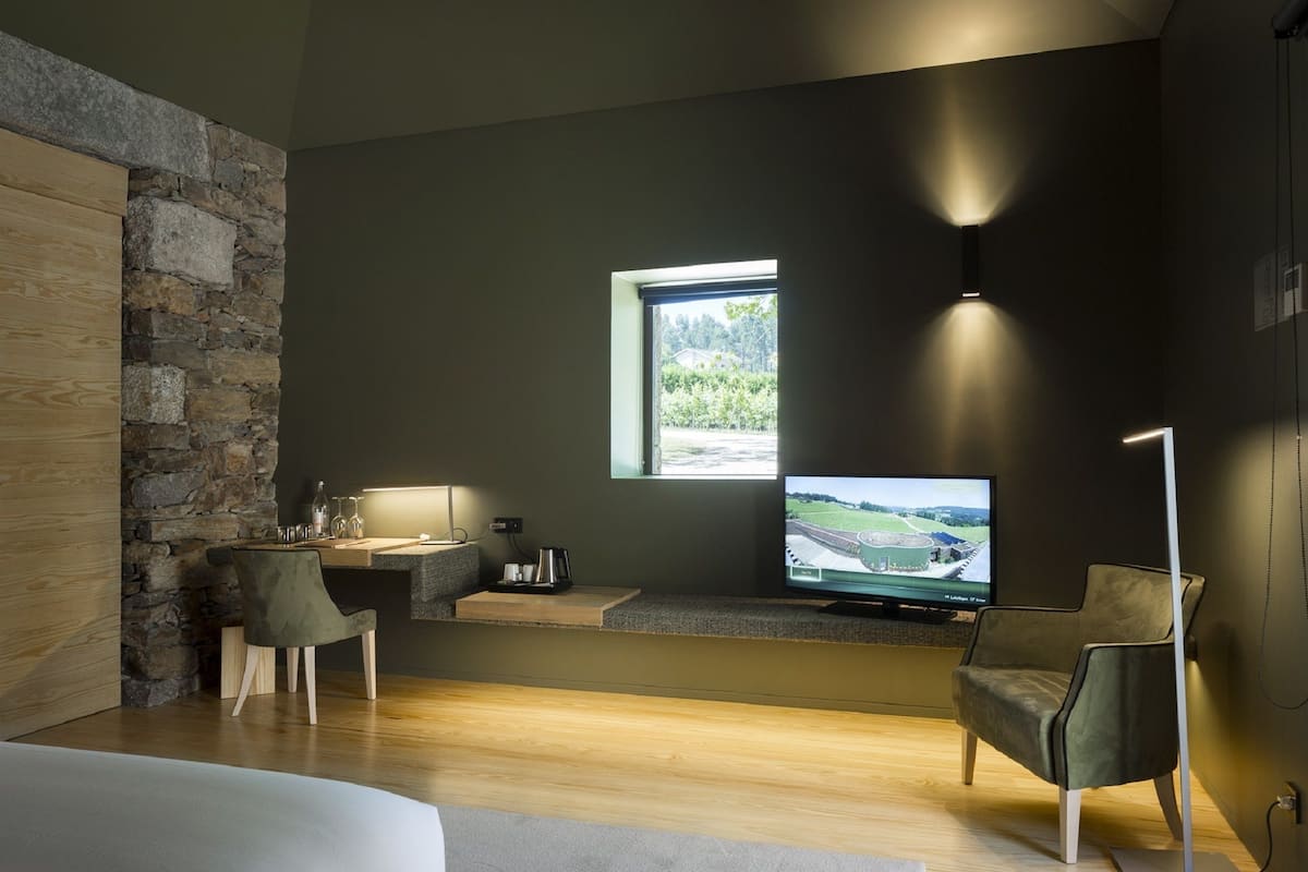 A modern hotel room with a stone accent wall, a large bed, a desk with a computer, a TV, and a view of a green landscape.