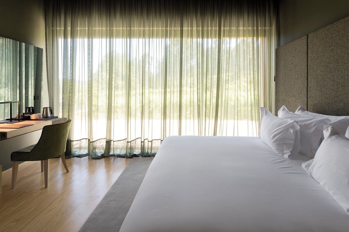 A hotel room with a large bed, a desk with a chair, and a large window with sheer curtains.