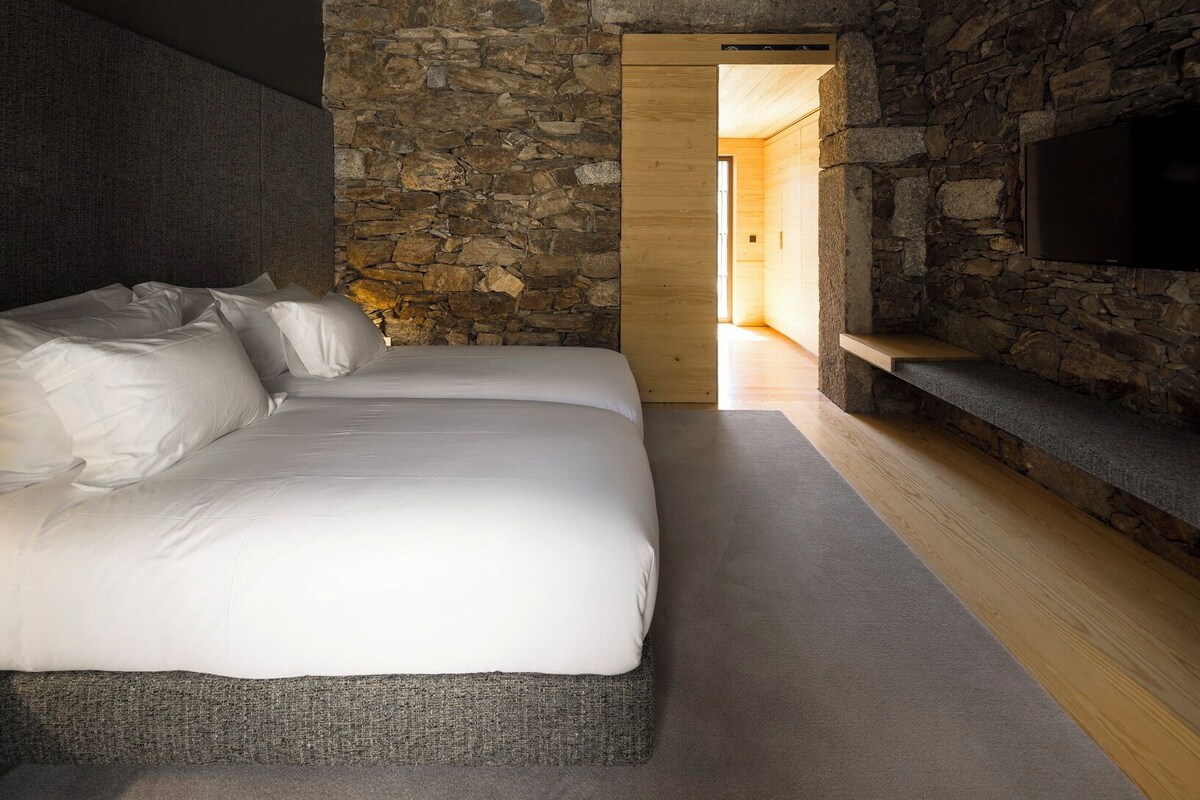 A bedroom with a stone wall, a bed with white bedding, and a television mounted on the wall.