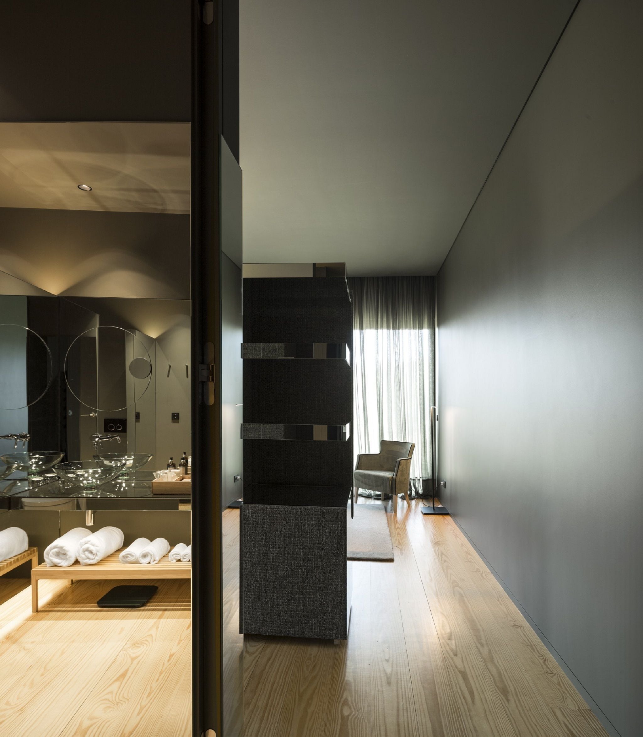 Un baño moderno con ducha envuelta en vidrio, tocador de madera y un rincón de descanso con una silla y una mesita.