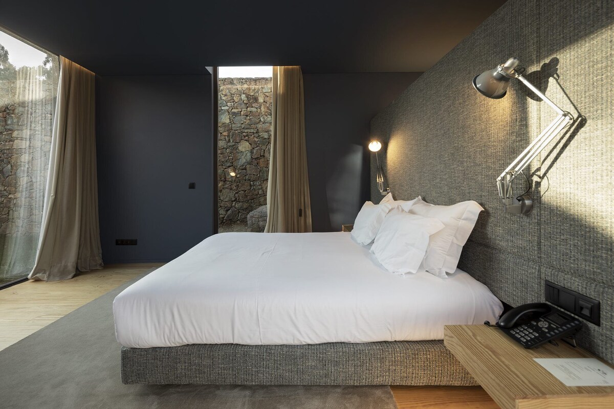 A modern bedroom with a large bed, a stone wall, and a view through a window.