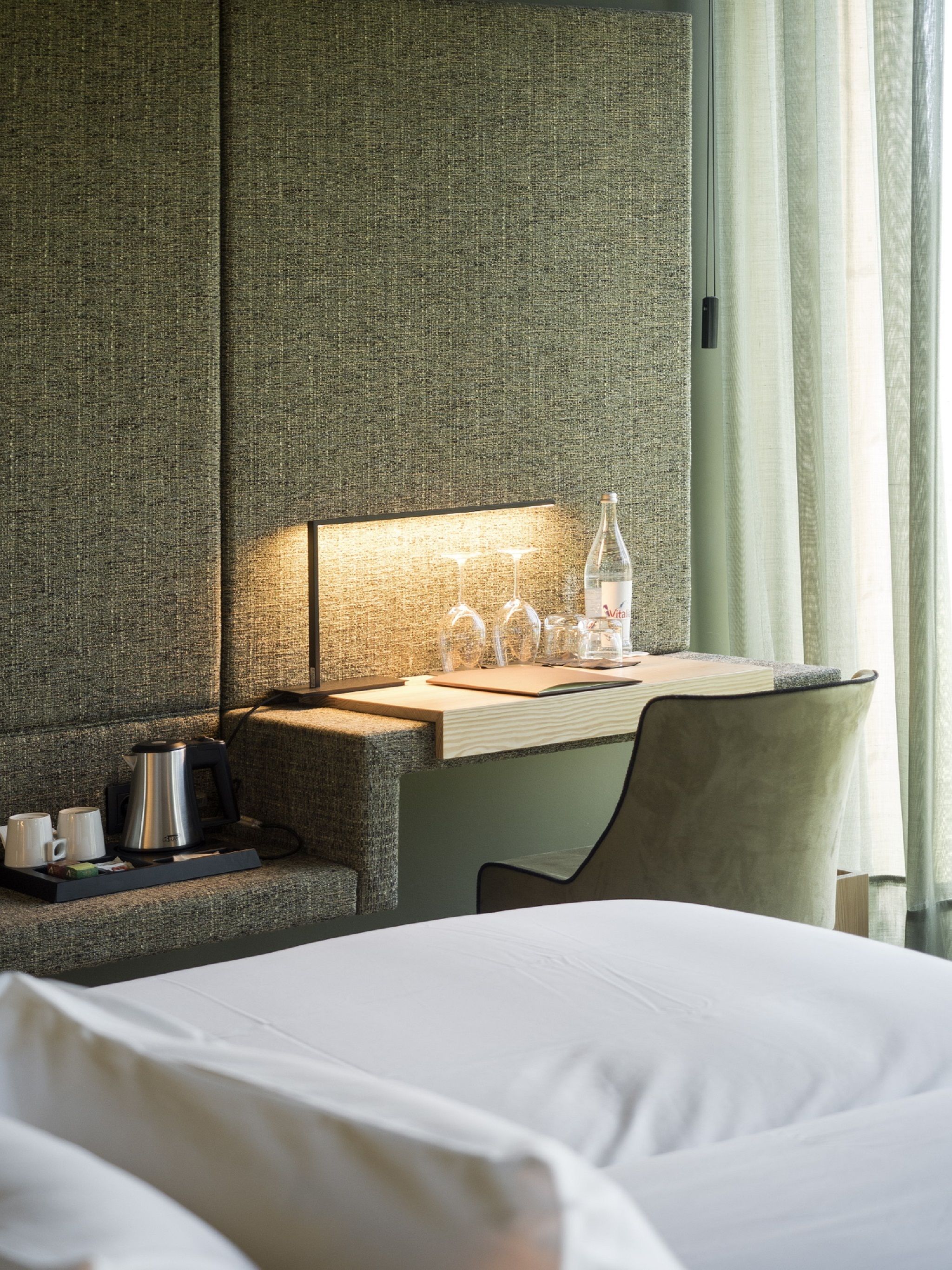 A hotel room with a bed, a desk with a lamp, a coffee maker, and a bottle of water.