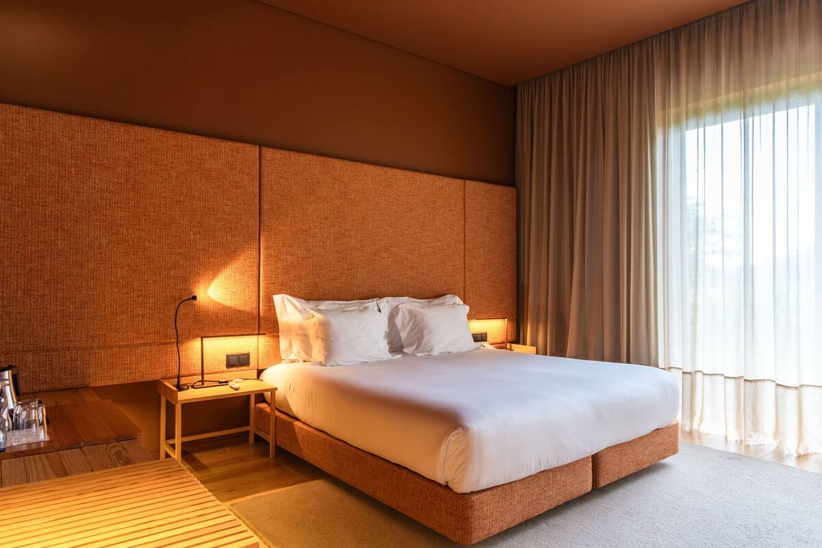 A modern hotel room with a large bed, a bedside table with a lamp, and a view of the outdoors through sheer curtains.