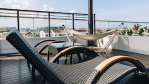 A rooftop terrace with wicker lounge chairs, a hammock, and a glass balustrade overlooking a cityscape.