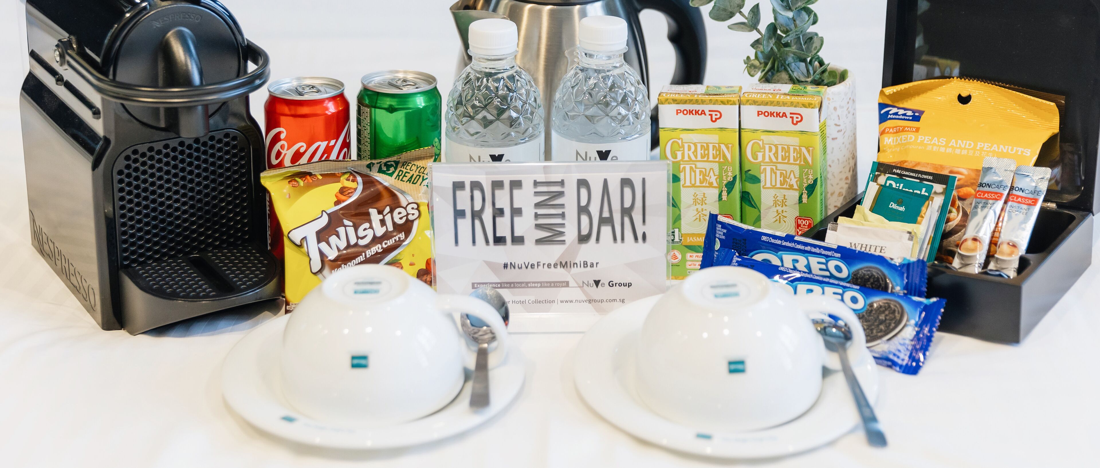 A breakfast setup with a Nespresso machine, two cups, a sign reading "FREE MINI BAR", and various snacks and drinks.