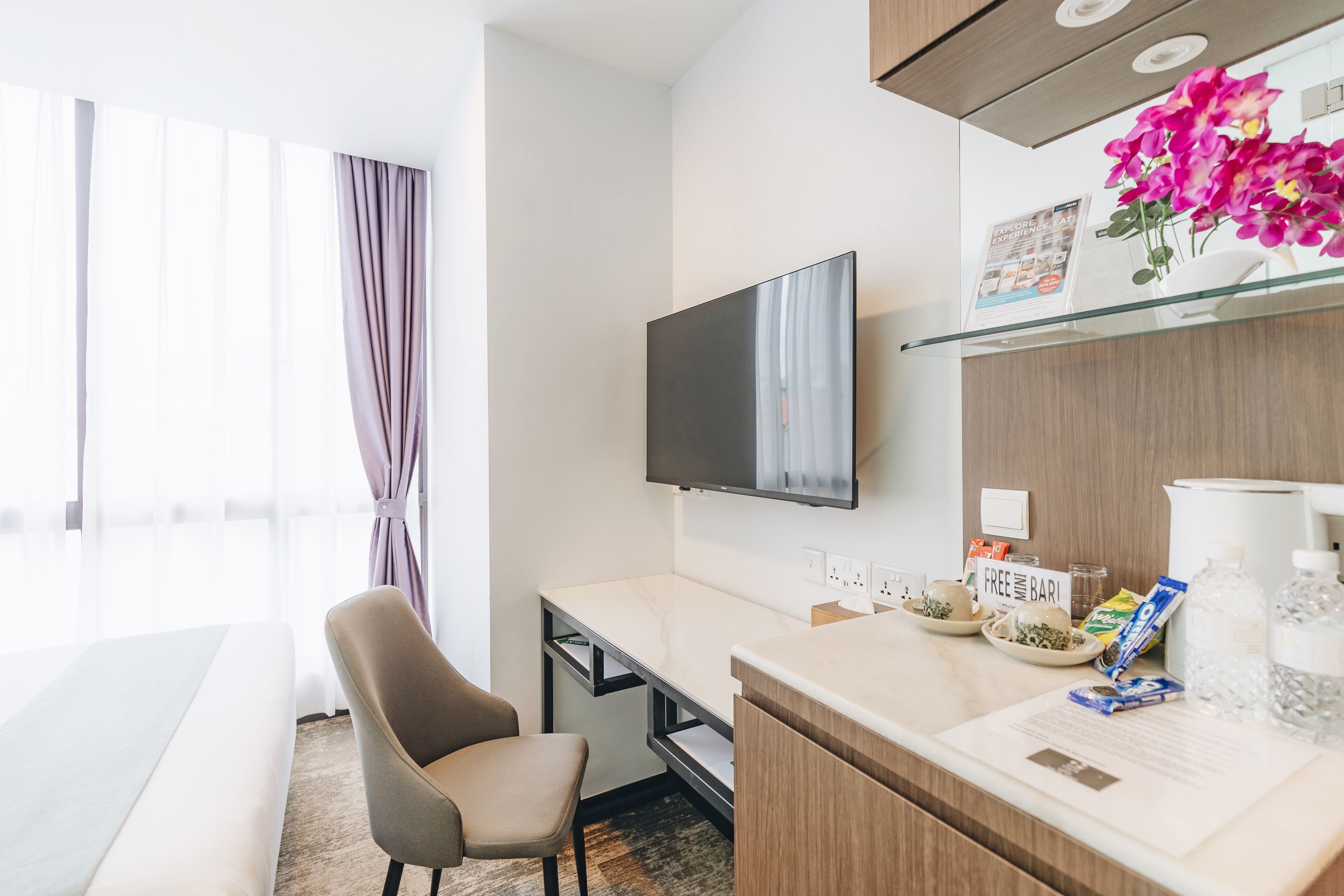 Habitación de hotel con cama, escritorio, televisor, mesita de vidrio con cafetera, cartel de 'Wi-Fi gratuito' y ventana con cortinas.