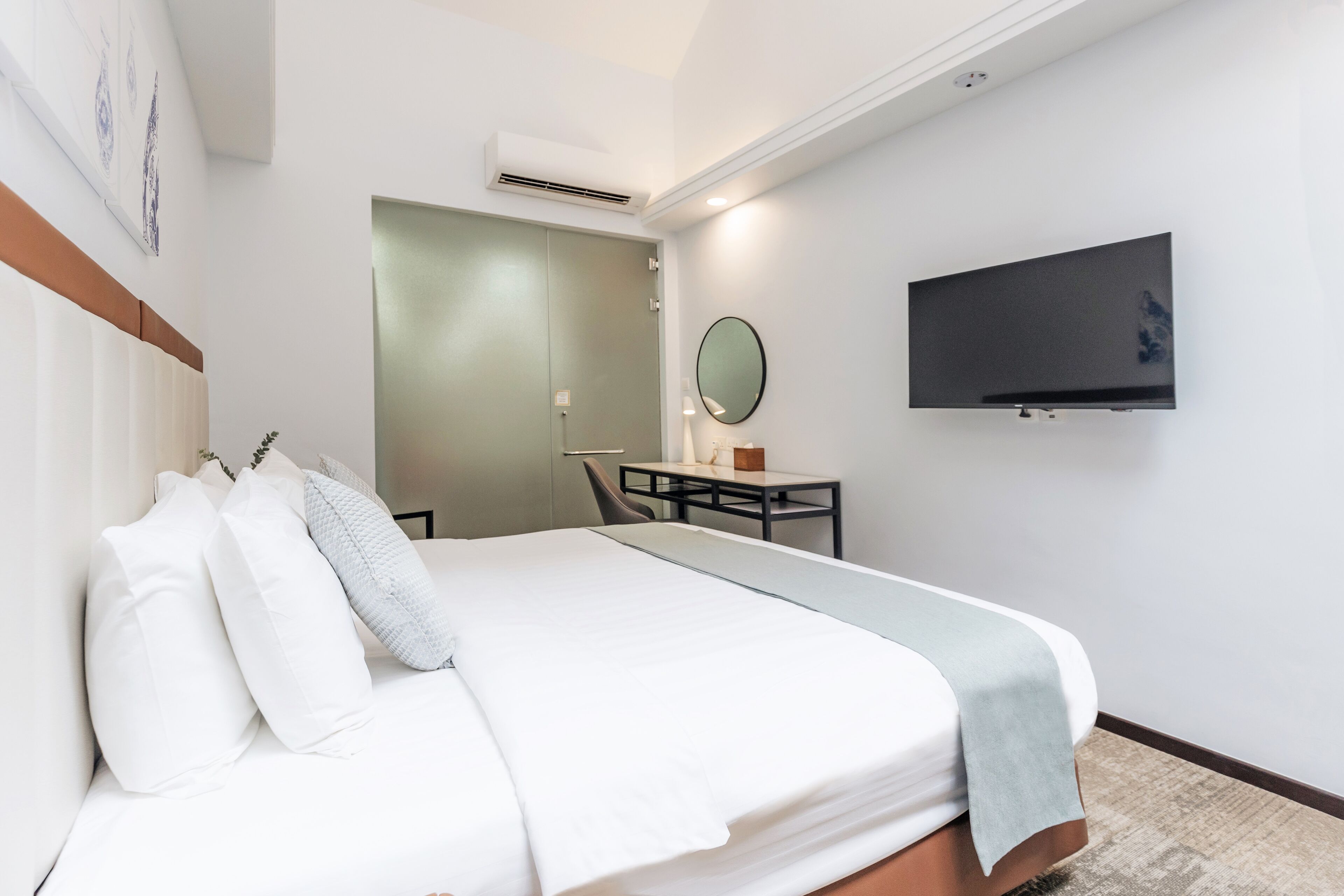 A hotel room with a bed, a desk, a chair, a TV, and an air conditioning unit.