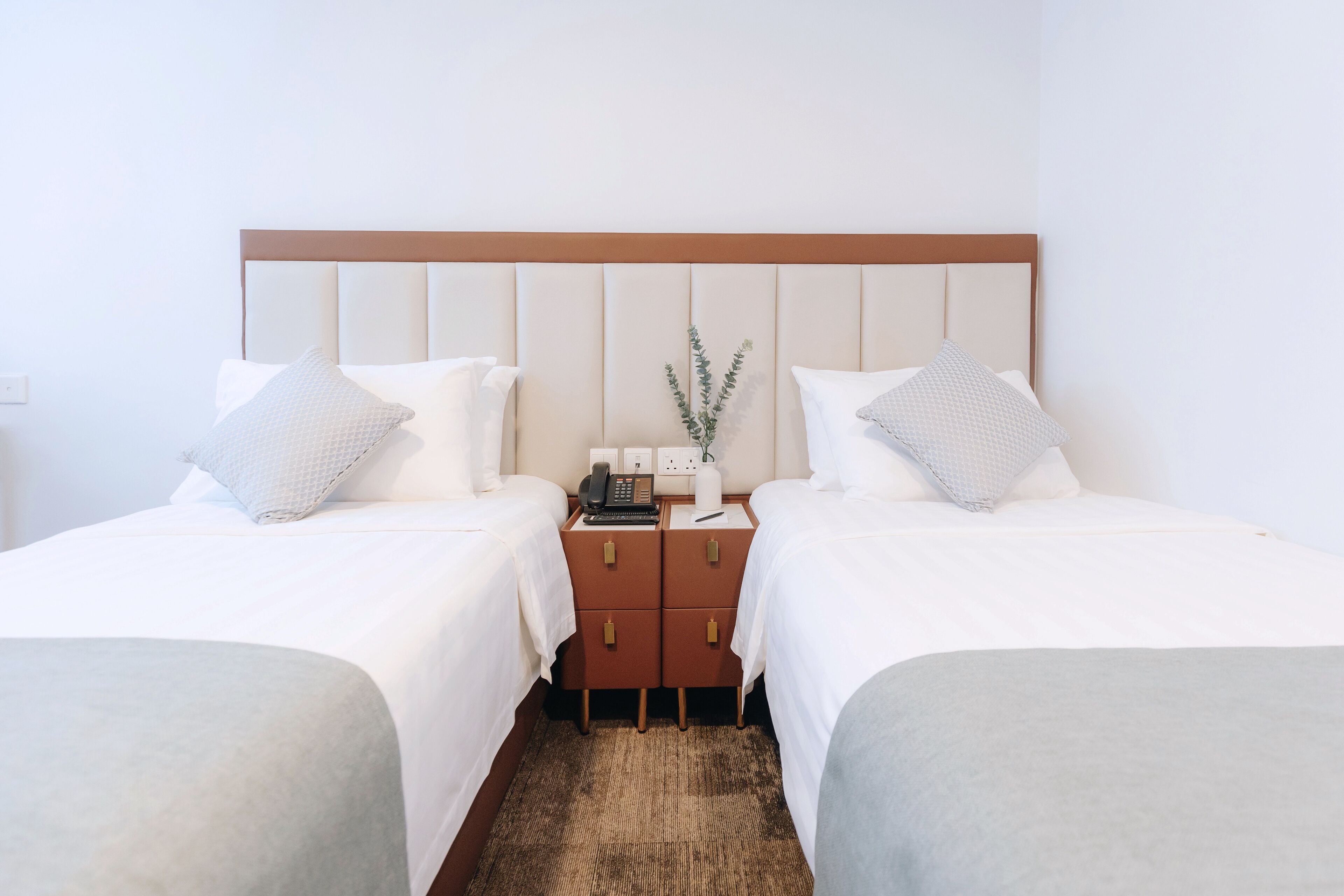 Habitación de hotel con dos camas, un cabecero de madera, una mesita de noche con un teléfono y un jarrón con una planta, sobre una alfombra marrón.