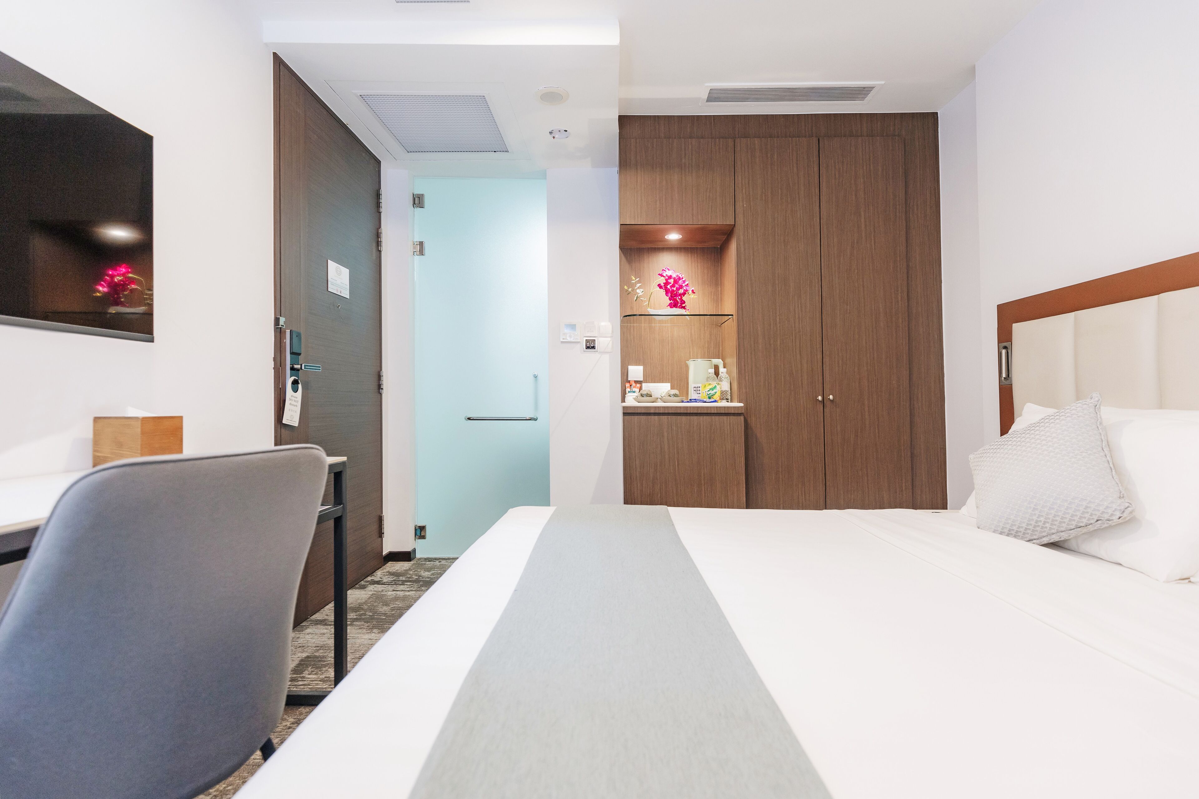 A hotel room with a bed, a desk, a chair, a wardrobe, and a small shelf with decorative items.