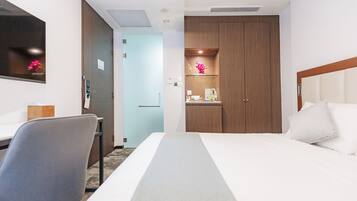 A hotel room with a bed, a desk, a chair, a wardrobe, and a small shelf with decorative items.