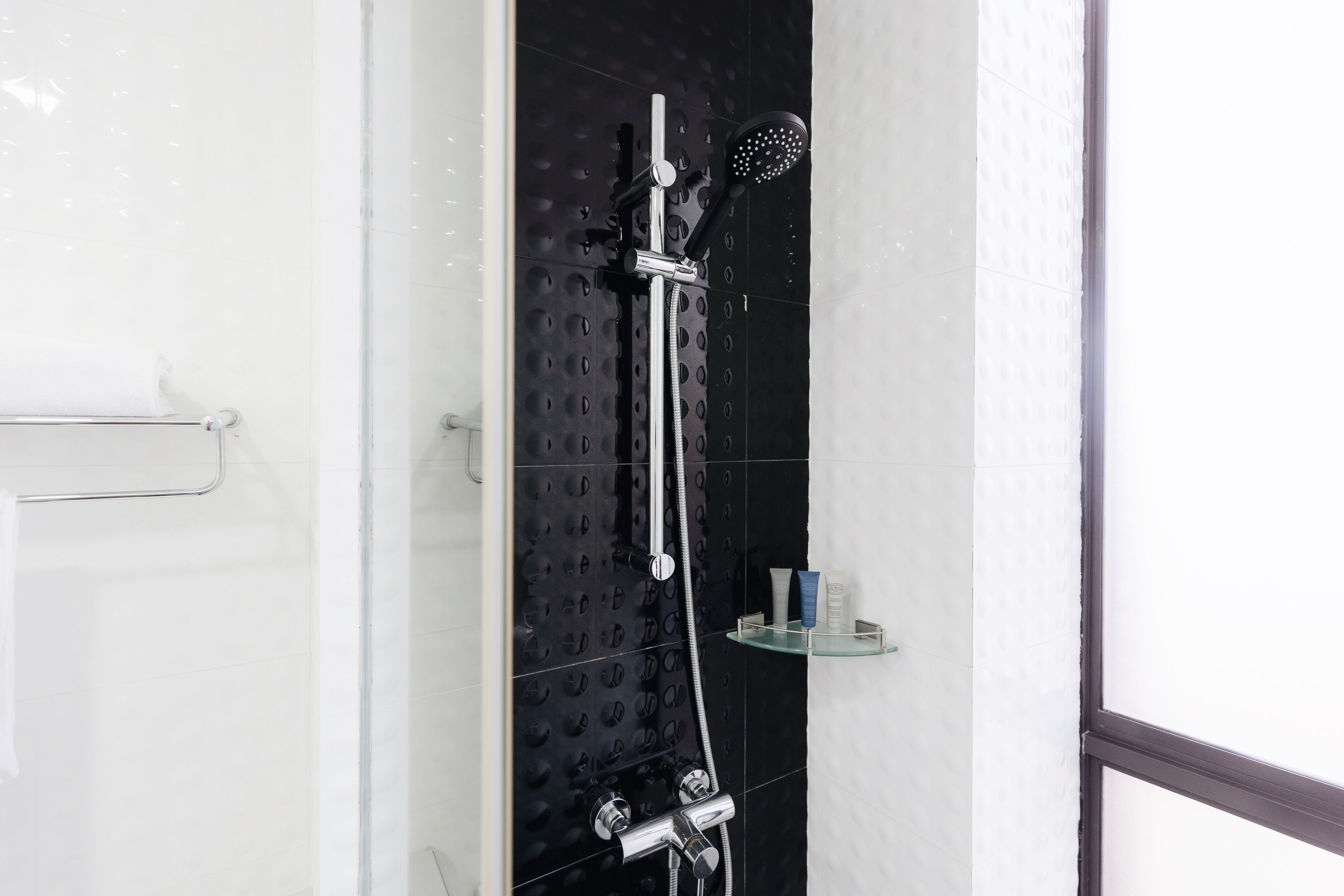 A shower with a handheld showerhead and a wall-mounted rainhead, a glass shelf with toiletries, and a towel rack.
