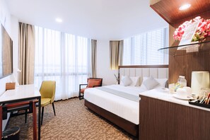 A hotel room with a large bed, a desk, and a chair. There is a window with curtains, a bedside table, and a nightstand. - Santa Grand Hotel East Coast, a NuVe Group Collection (Singapore)