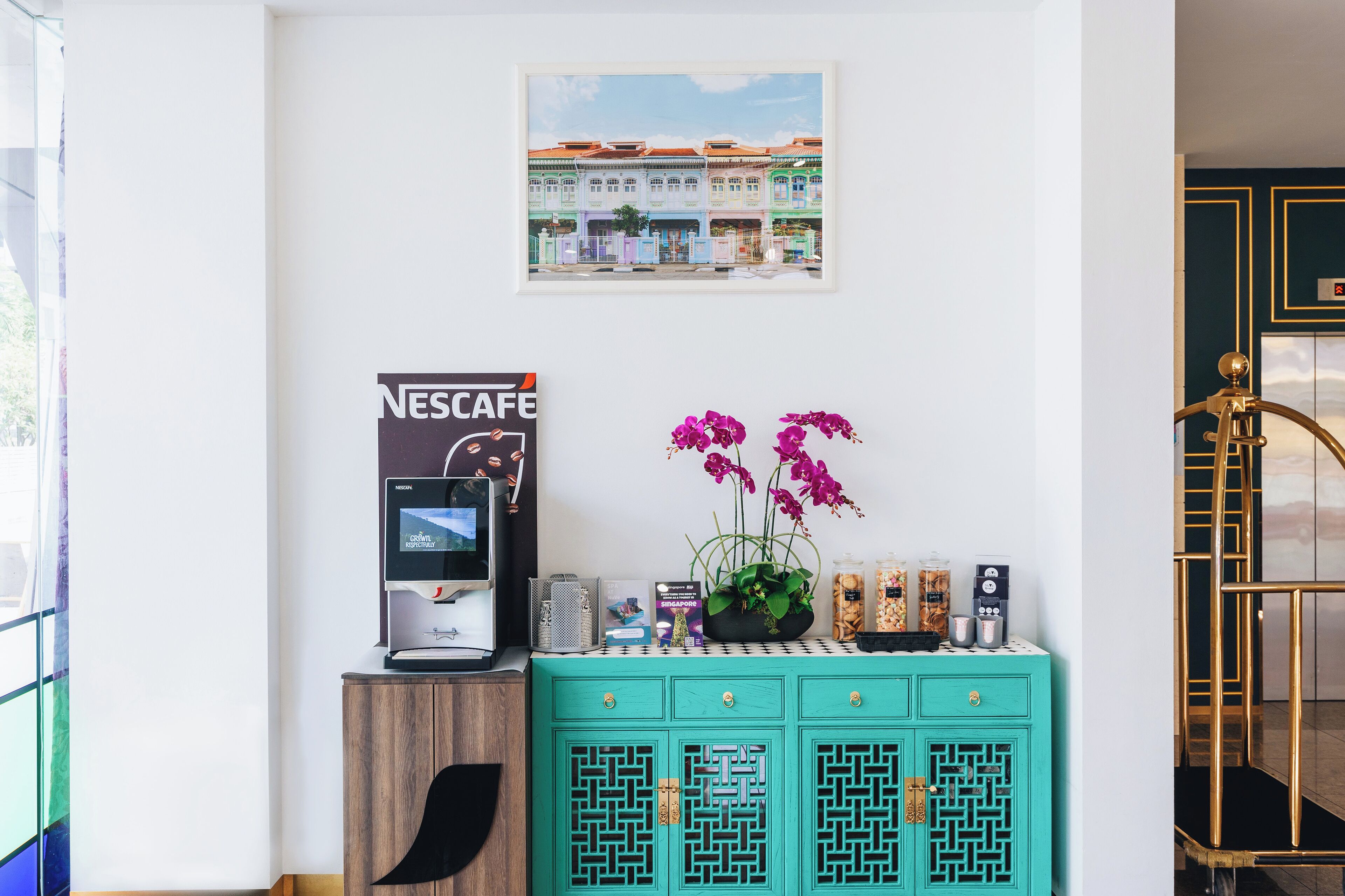 Una máquina de café Nescafé, una orquídea en maceta y un cuadro con una imagen de un edificio sobre un mueble color turquesa.