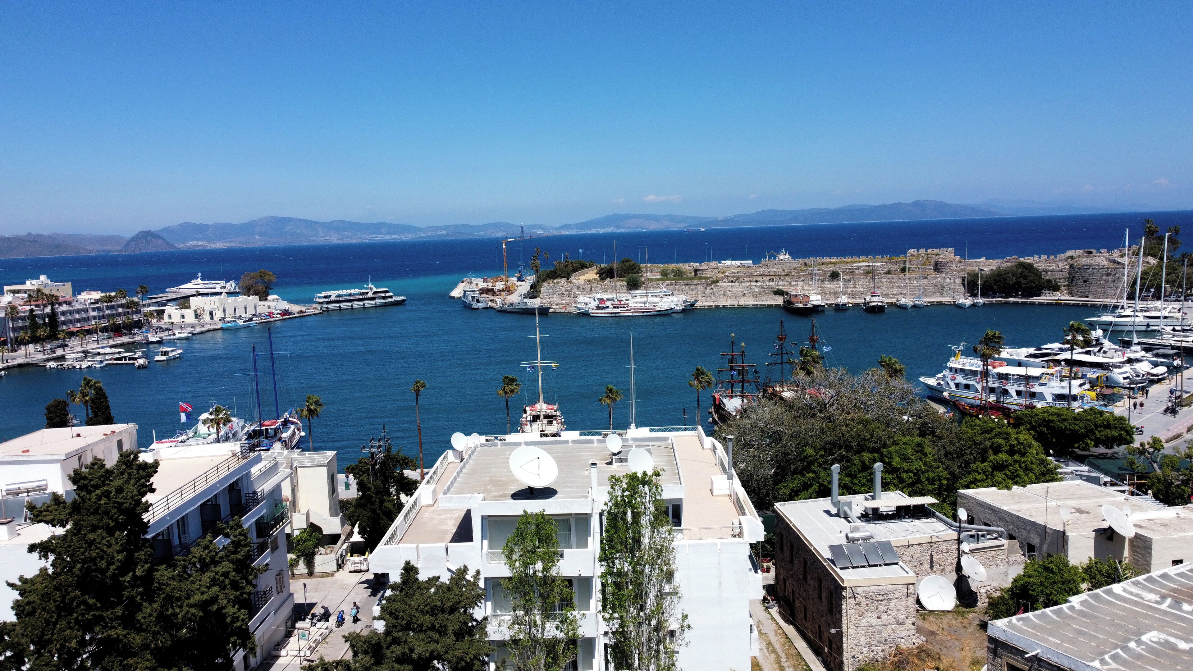 Un port de plaisance situé sur la côte, avec des bateaux amarrés, un fort historique et des immeubles résidentiels équipés d’antennes paraboliques.
